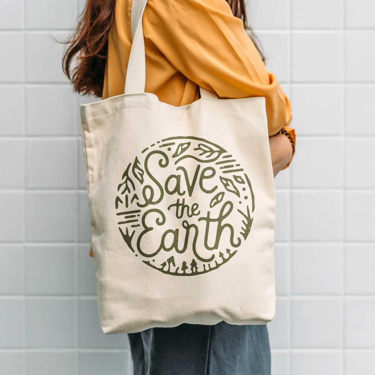 Person holding a tote bag with 'Save the Earth' text against a tiled wall.